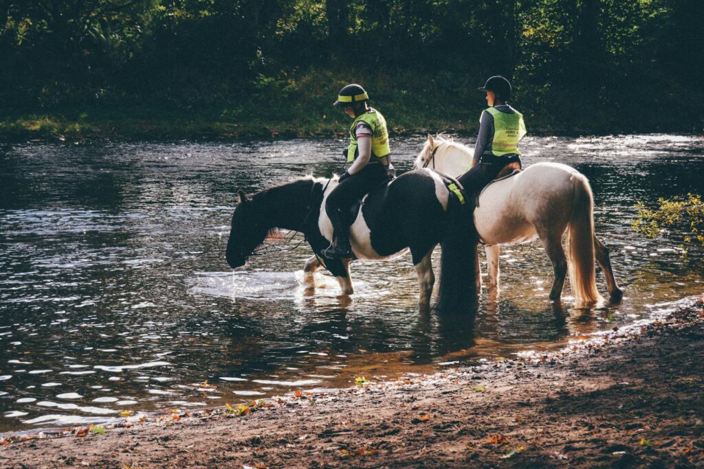 Two equestrians ride horses by a tranquil river in Scotland, showcasing the beauty of nature.