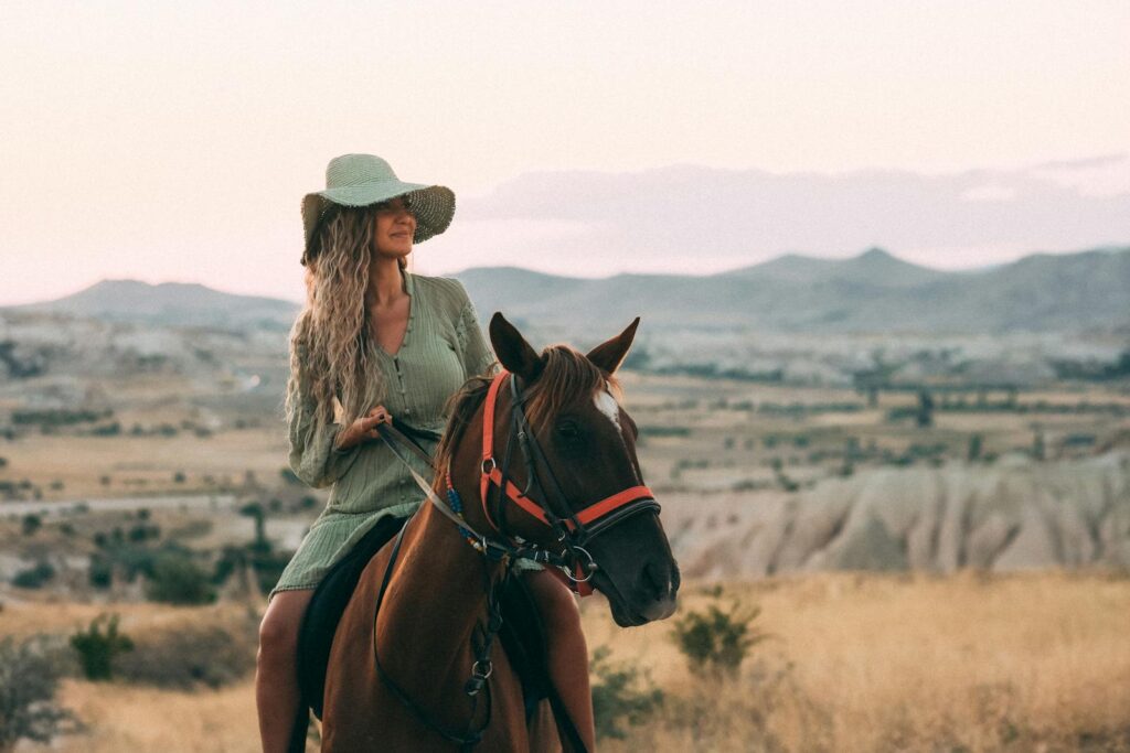 A woman with long blonde hair and a sun hat rides a horse in a picturesque rural landscape.