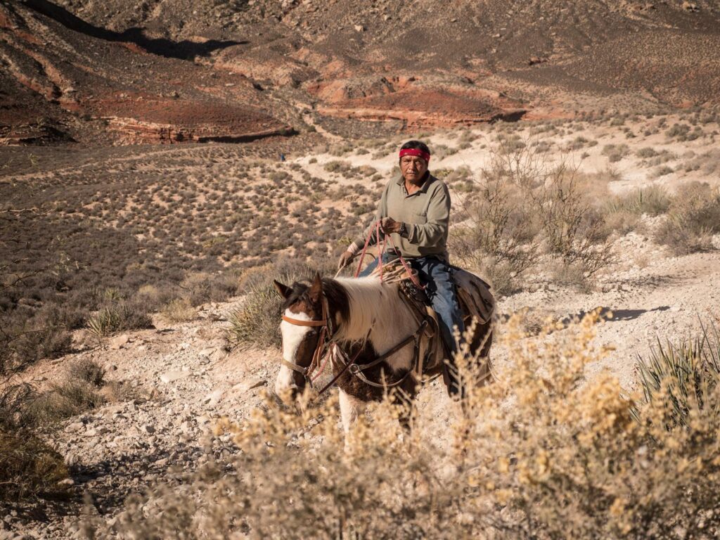 Indigenous man riding a horse through a rugged desert valley under the summer sun.