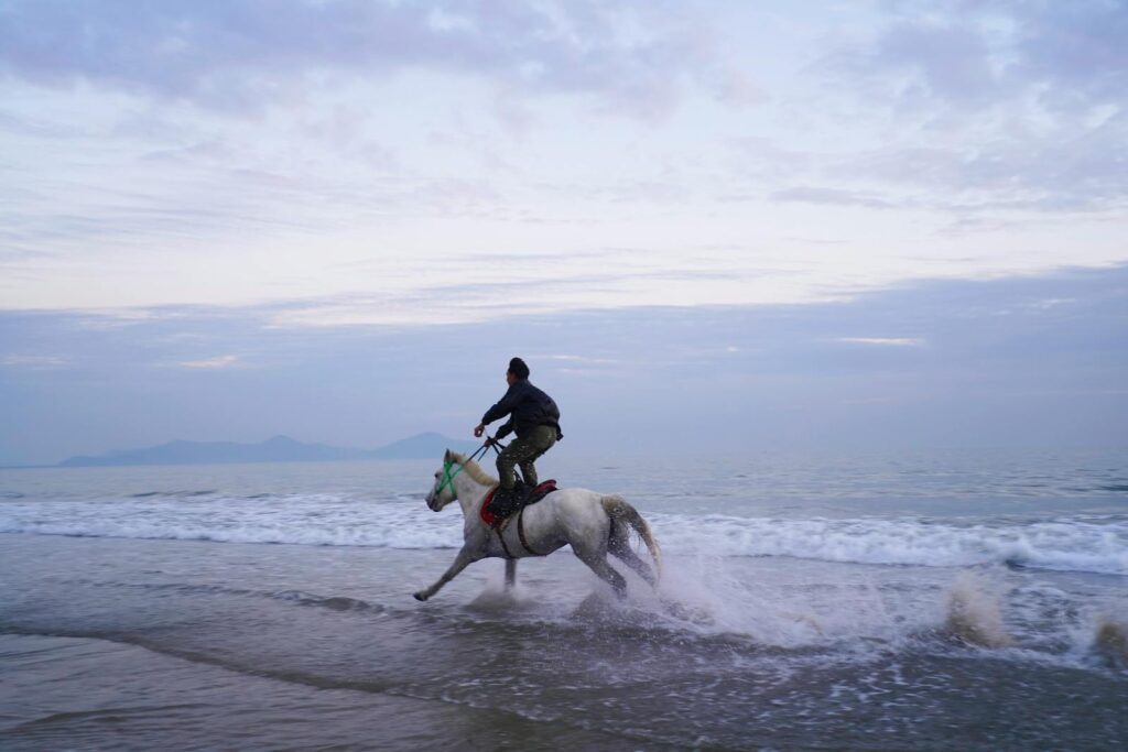 A man rides a galloping horse along a serene beach, with waves splashing under a cloudy sky.