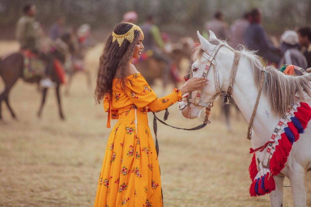 A woman in a vibrant dress stands tenderly touching a decorated white horse in a field.