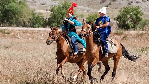 Two people in traditional Kyrgyz clothing riding horses across a grassy field
