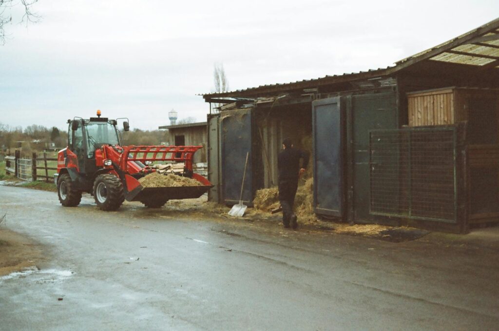 A farmer working near a barn with a tractor loading hay, under an overcast sky.