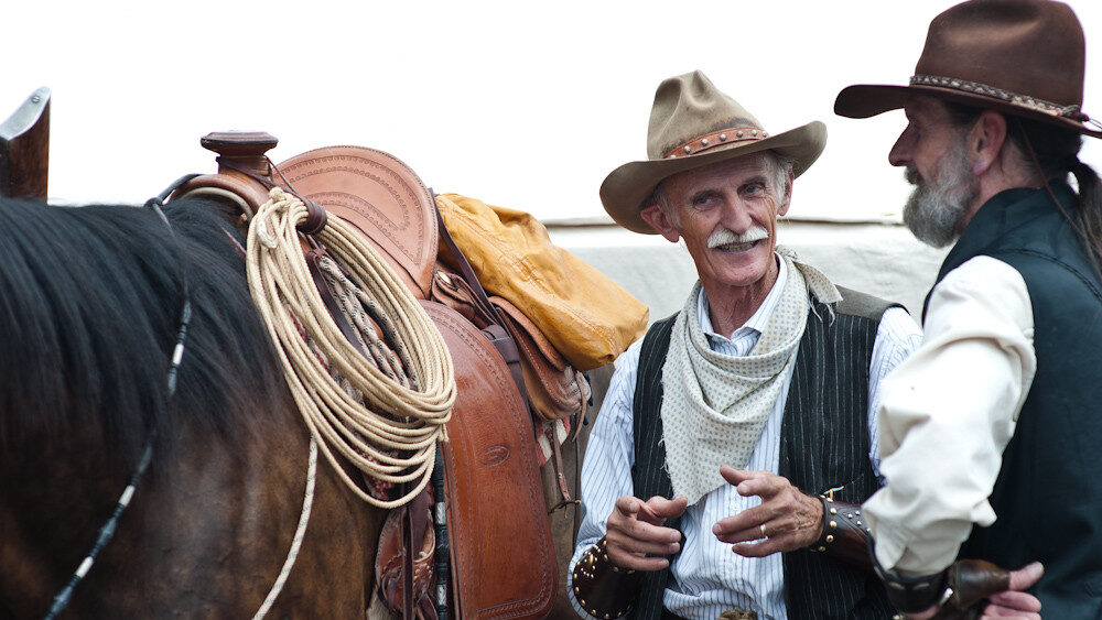Two ranch hands are talking to each other regarding the horse beside them