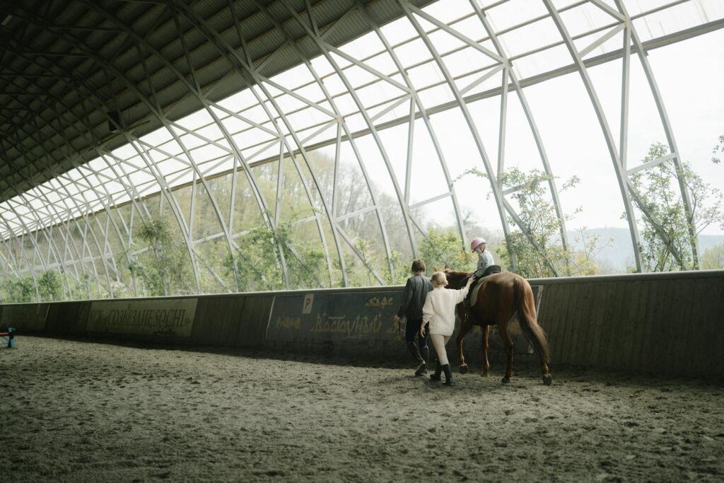Child learning horseback riding in an indoor equestrian arena with guidance from instructors.
