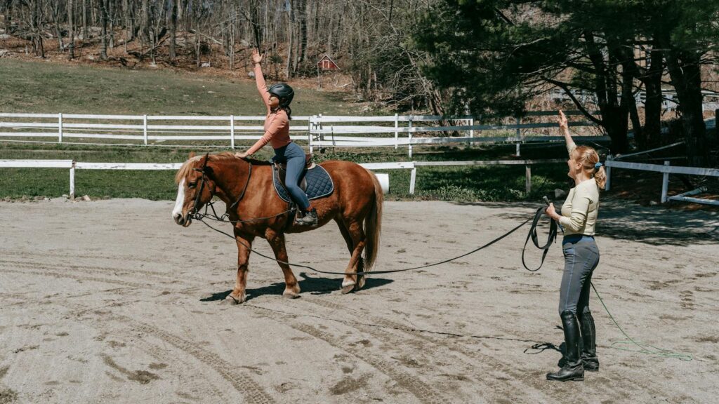 Woman training rider on horse in a sunny outdoor arena with natural surroundings.