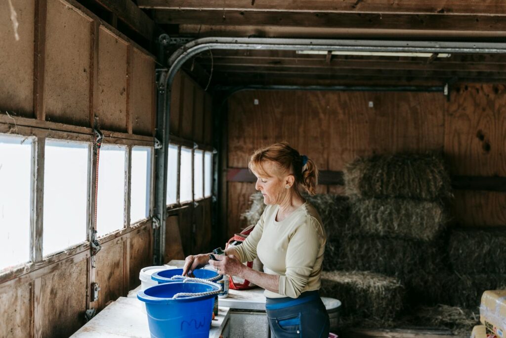 A woman arranging blue buckets in a barn filled with hay bales, standing by a window.
