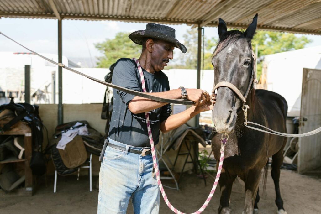 Man in cowboy hat grooming a horse inside a rustic stable