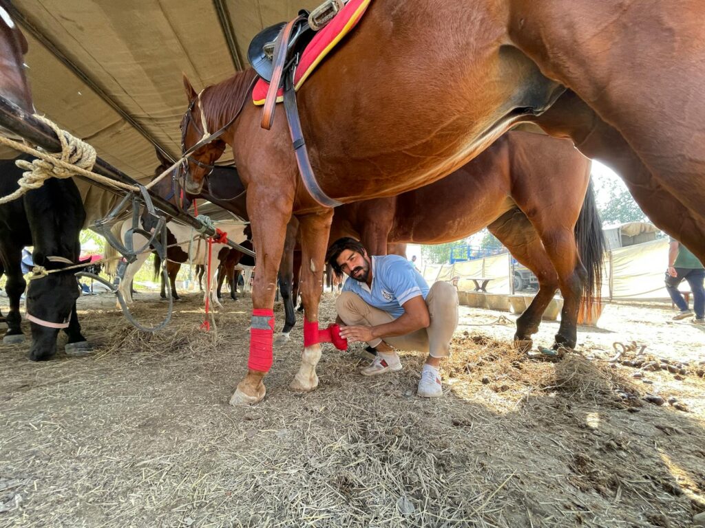 A man is wrapping a horse's leg with red tape at an equestrian event, surrounded by other horses.
