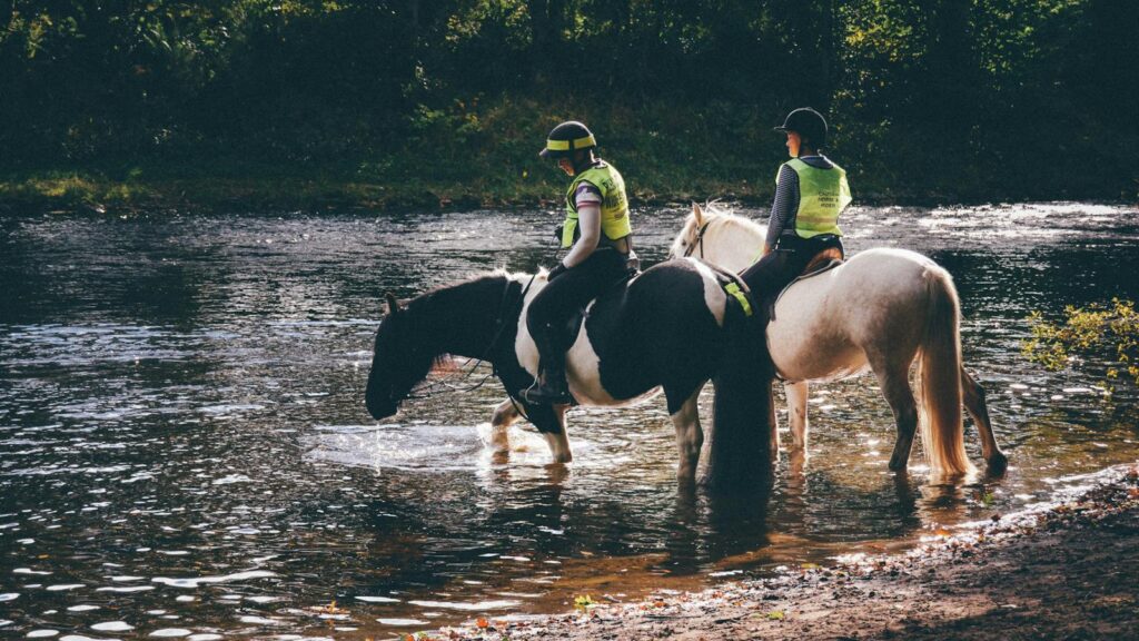 Two equestrians ride horses by a tranquil river in Scotland, showcasing the beauty of nature.