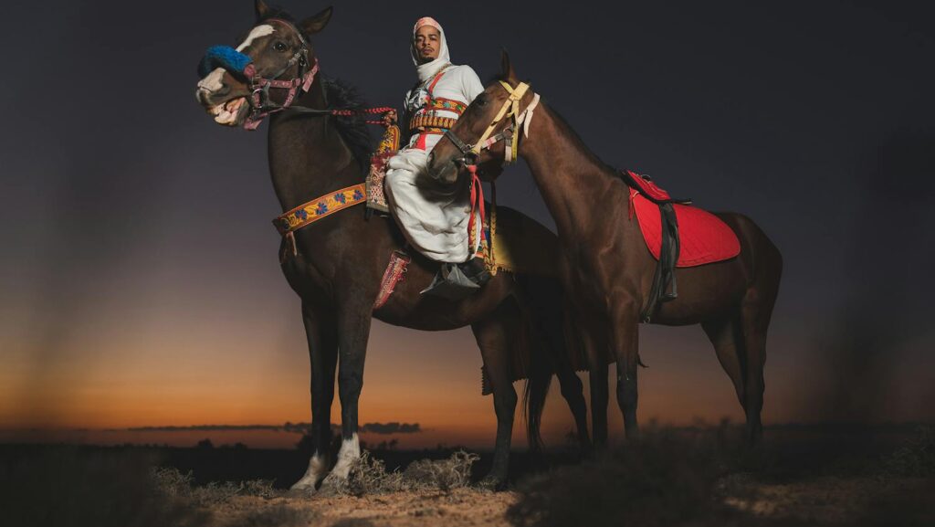 Man in traditional attire riding horses during a stunning Tunisian sunset.