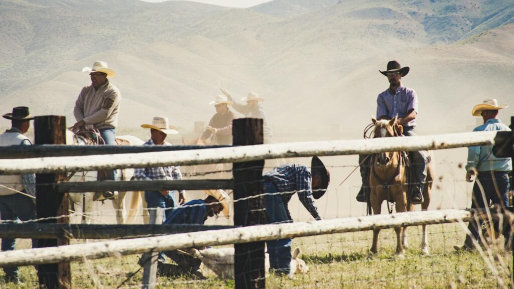 Rural landscape with cowboys on horseback wrangling cattle in a dusty field.