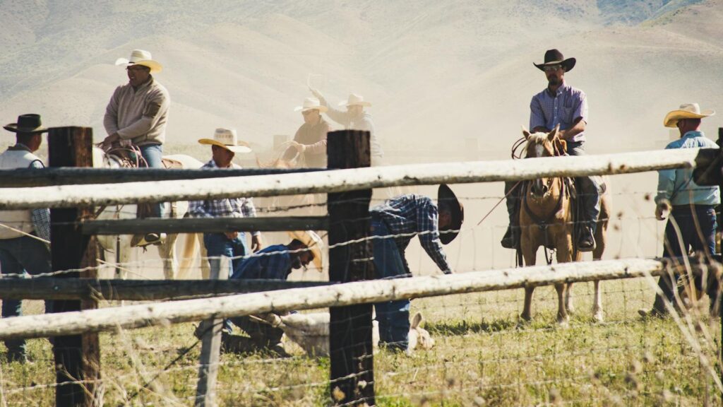 Rural landscape with cowboys on horseback wrangling cattle in a dusty field.