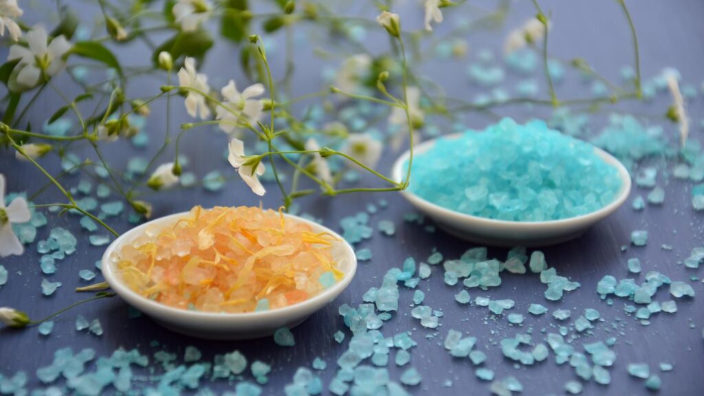 bath salts in dishes surrounded by delicate white flowers on a blue surface.