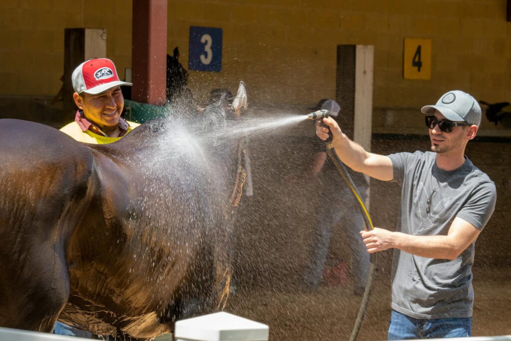 Two men using a water hose to wash a horse in an outdoor stable setting.