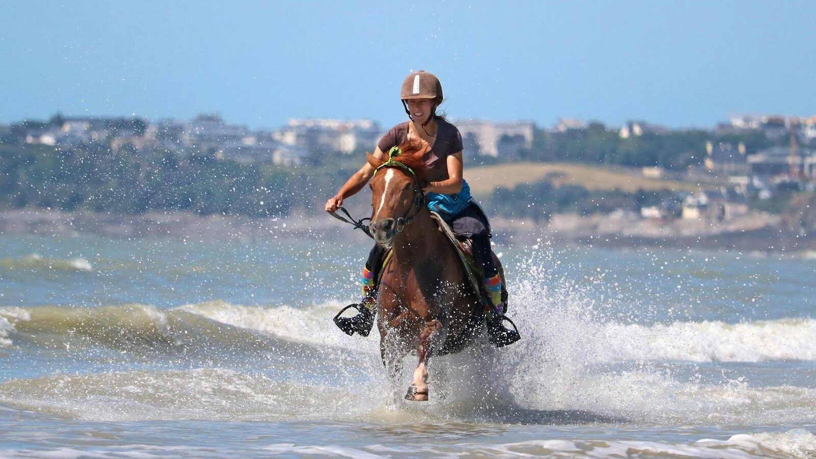 A woman rides a horse through ocean waves on Granville beach, Normandie, France.