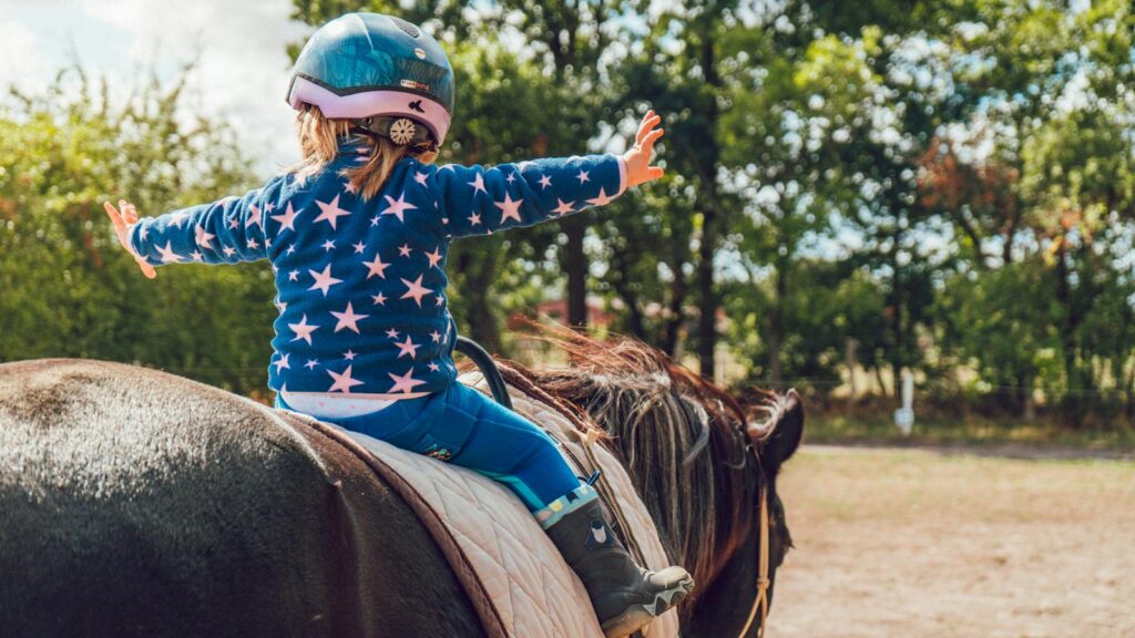 A child confidently rides a horse outdoors, wearing a helmet and star-patterned jacket.