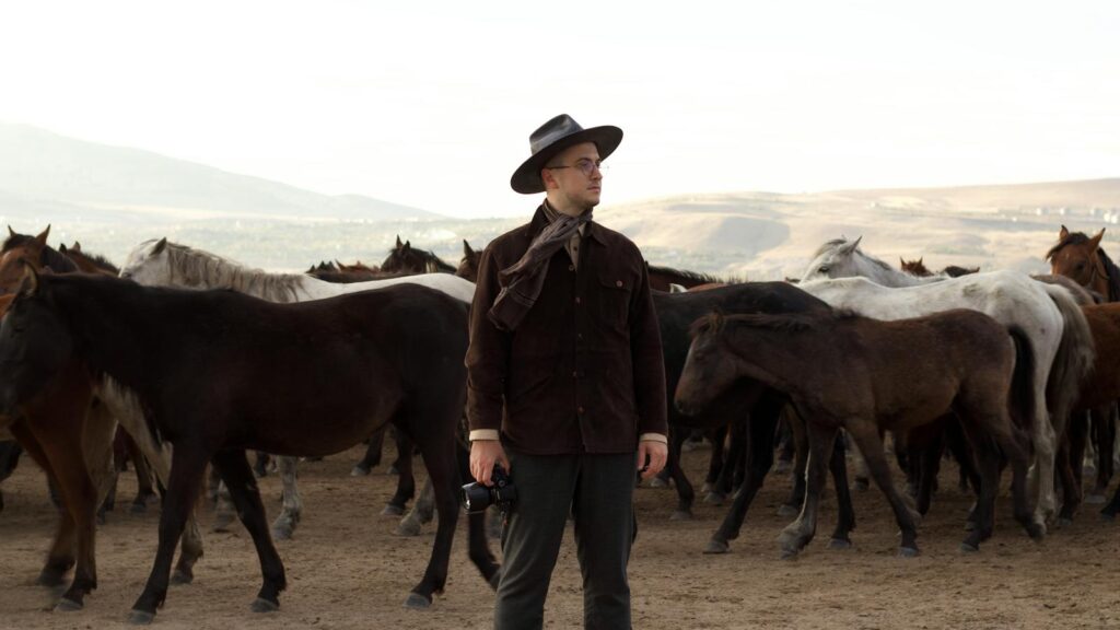A man with a camera stands among a herd of wild horses in a vast, open landscape.