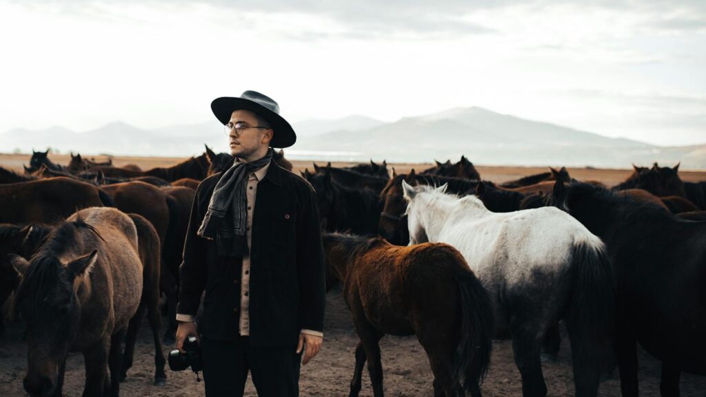 Man in cowboy hat among a herd of horses in a tranquil, open landscape.