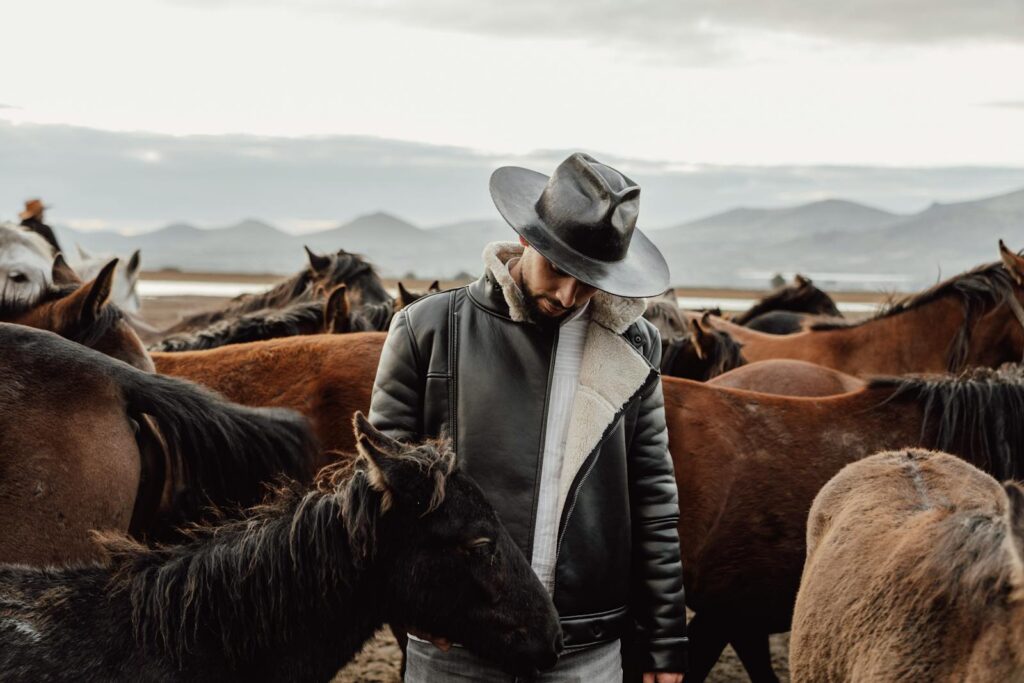 A cowboy in a leather jacket and hat stands among a herd of horses in a scenic outdoor field with mountains in the background.