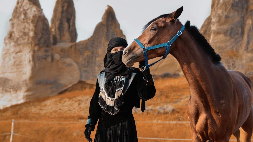 A person in traditional attire stands with a horse amidst striking rock formations in a desert setting.