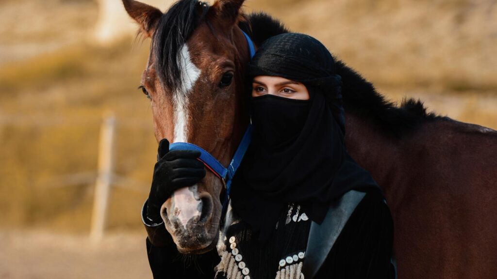 A woman in traditional clothing poses with a horse in a desert landscape.