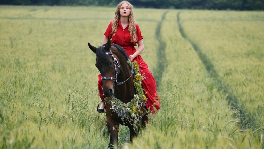 A blonde woman in a red dress riding a horse through a rural field.