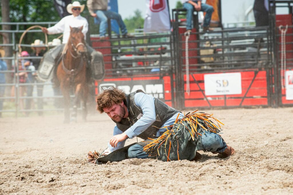 A cowboy falls in a rodeo competition with a horse in the background. Action shot.