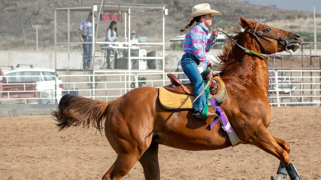 A young cowgirl rides a horse during a rodeo competition at an outdoor arena.