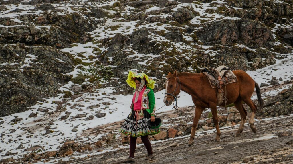 An Andean woman in traditional attire walks beside a horse in a snowy mountain landscape.