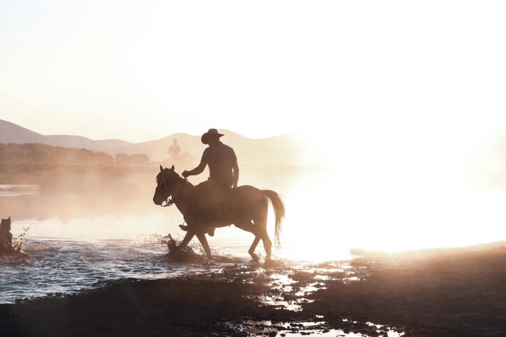 A cowboy riding a horse through water at sunset, creating a striking silhouette against a bright sky.