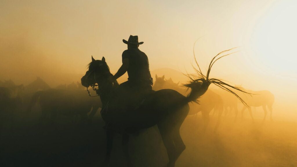 A cowboy riding a horse through a dusty sunrise, creating a dramatic silhouette.