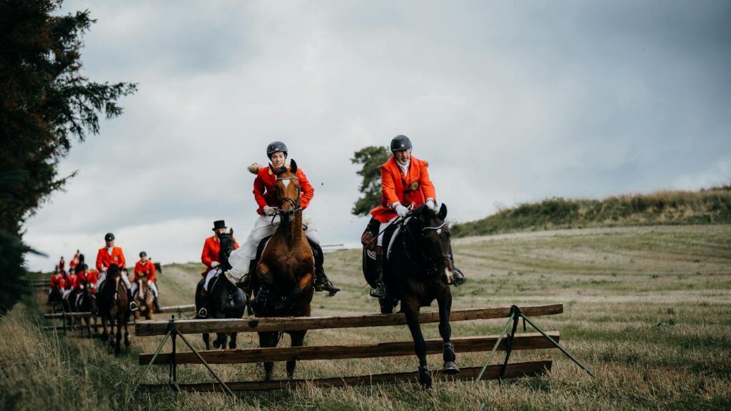 Horse riders in red jackets jumping over fences in a traditional fox hunting event on a rural landscape.