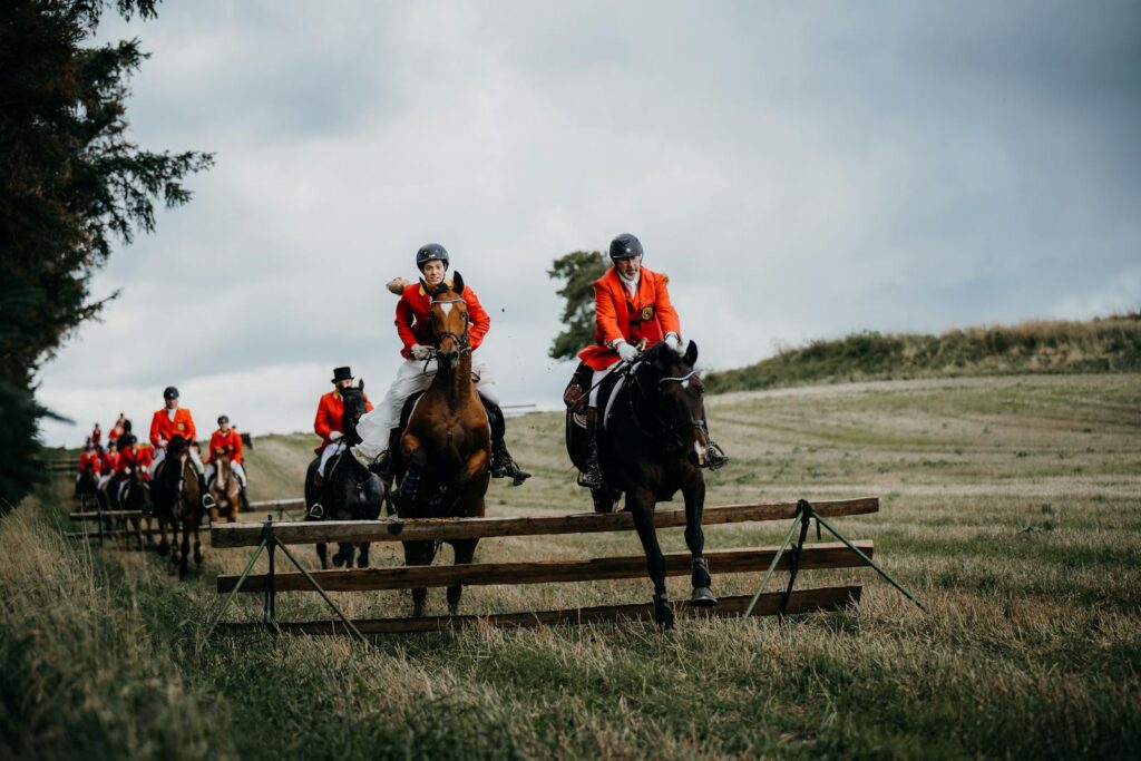 Horse riders in red jackets jumping over fences in a traditional fox hunting event on a rural landscape.