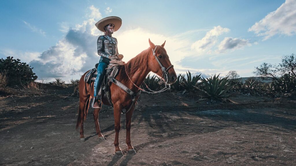A man in traditional Mexican attire riding a horse in a rural landscape under a bright blue sky.