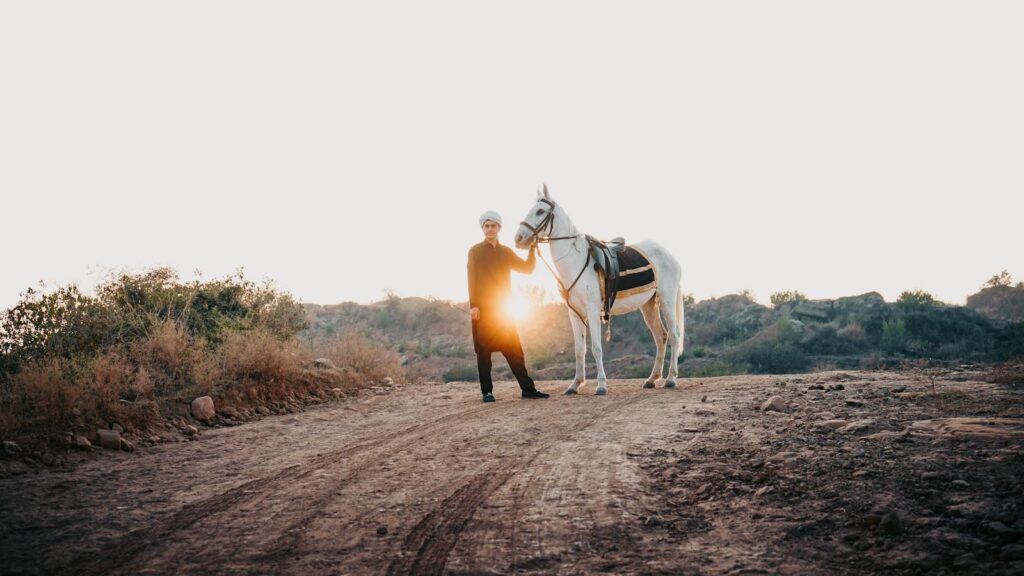 A man in traditional attire with a horse, set against a scenic countryside sunset.