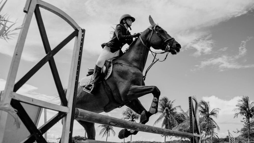 A woman riding a horse mid-jump during an equestrian event in a tropical outdoor setting.