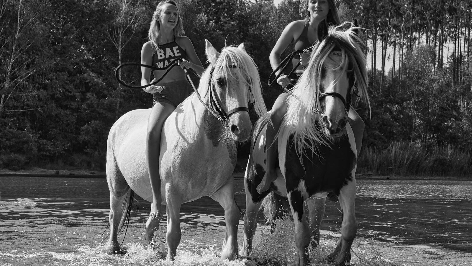 Two women riding horses through a stream in a summer forest setting, black and white photo.