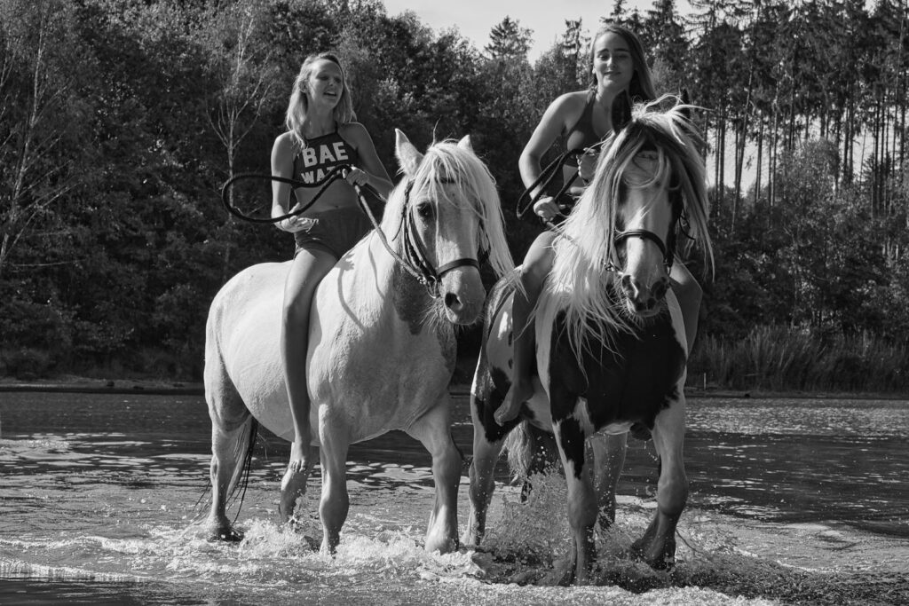 Two women riding horses through a stream in a summer forest setting, black and white photo.