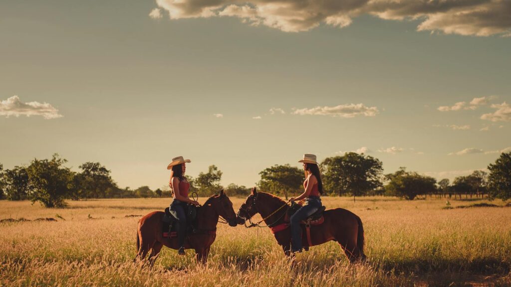 Two women horseback riding in a sunny Brazilian field during sunset, showcasing nature's serenity.