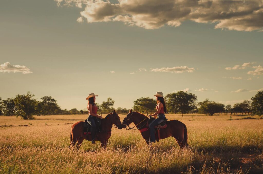 Two women horseback riding in a sunny Brazilian field during sunset, showcasing nature's serenity.