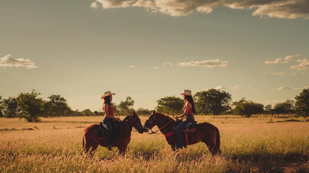 Two women horseback riding in a sunny Brazilian field during sunset, showcasing nature's serenity.