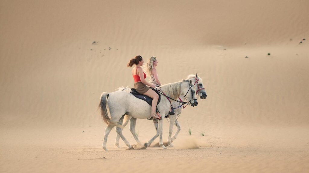 Two women riding horses through the dunes of Dubai's desert, capturing serene beauty and adventure.
