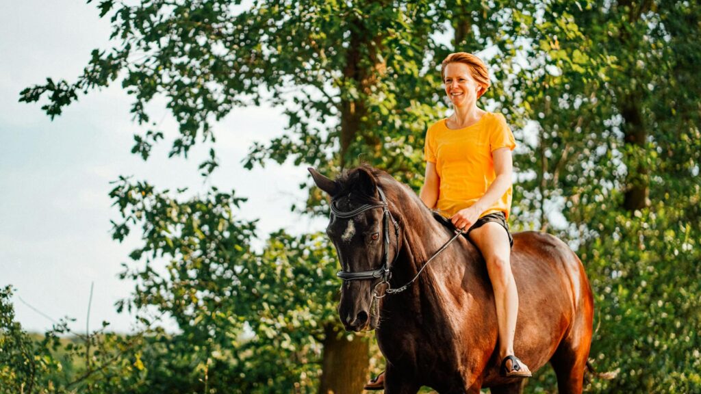 A woman in a yellow shirt enjoys horse riding through a sunny forest trail.