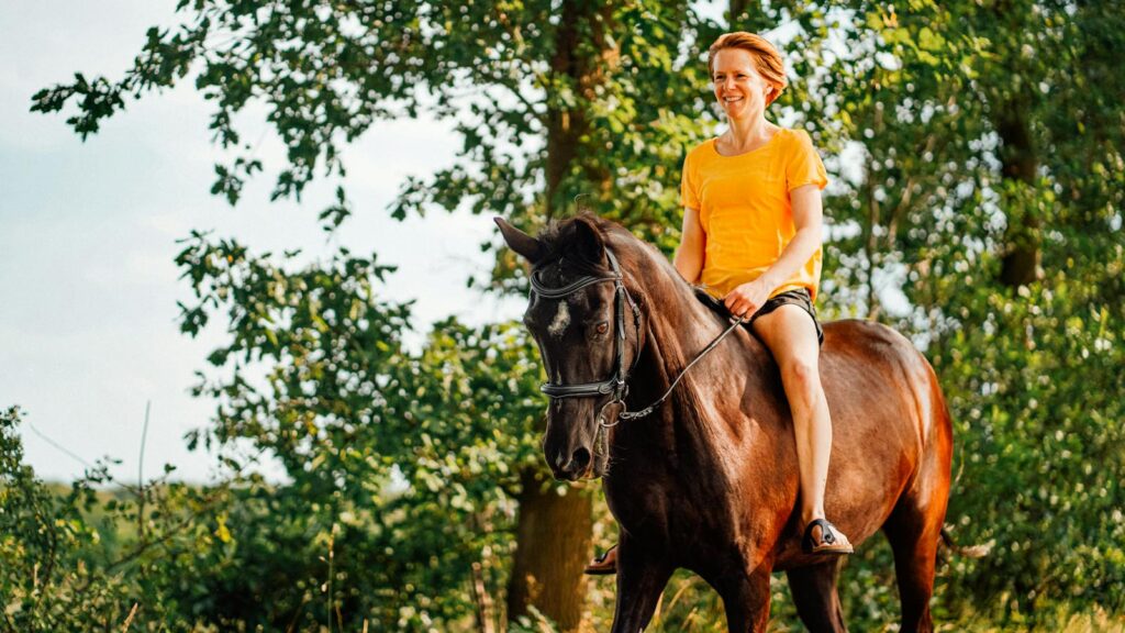 A woman in a yellow shirt enjoys horse riding through a sunny forest trail.