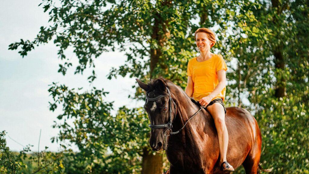 A woman in a yellow shirt enjoys horse riding through a sunny forest trail.