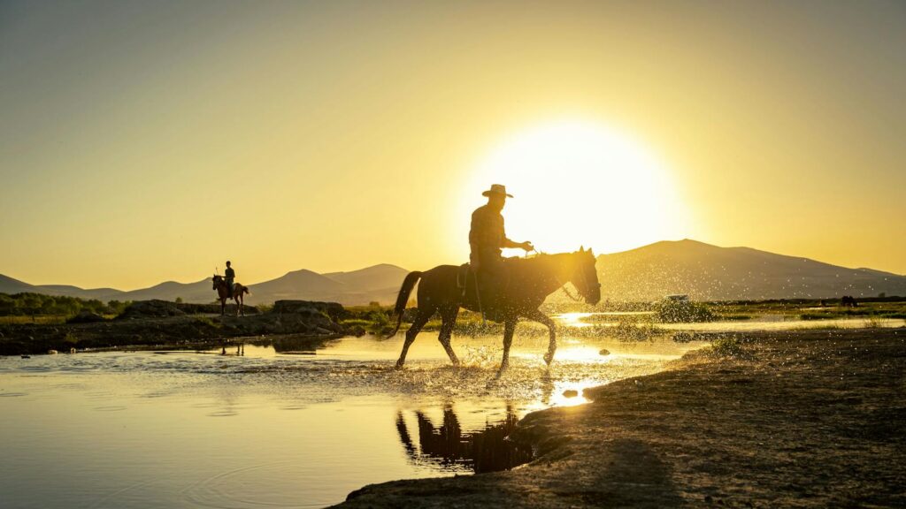 Silhouette of a cowboy on horseback crossing a water stream against a stunning sunset backdrop.