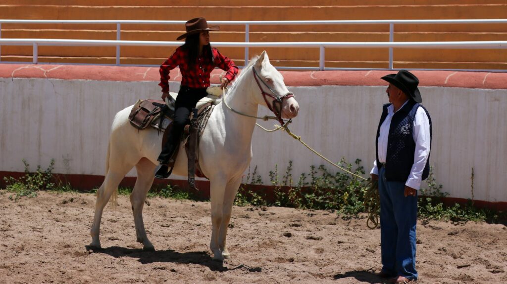 A woman in a plaid shirt on a horse practicing with a man in a cowboy hat in an outdoor arena.