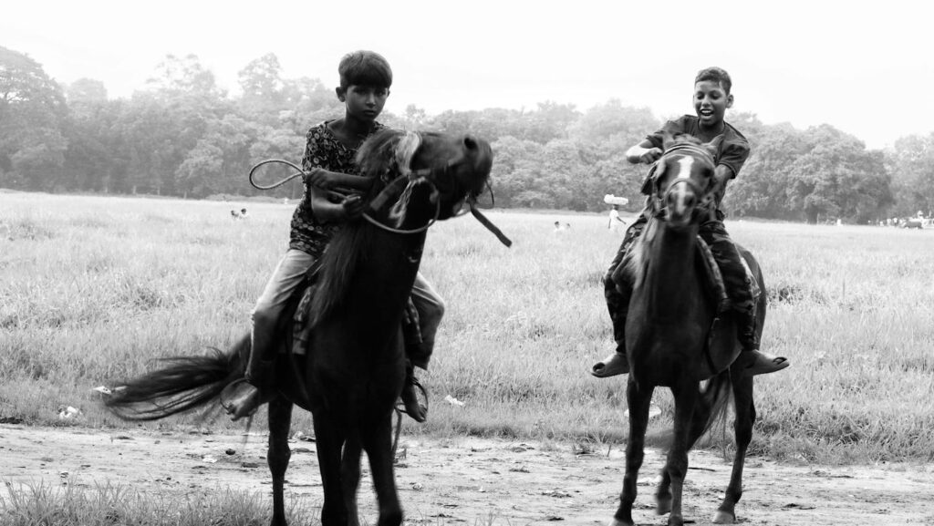 Two boys riding horses in an open field, captured in black and white.
