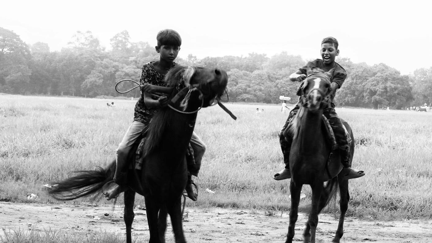 Two boys riding horses in an open field, captured in black and white.